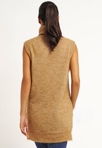 Pull sans manches beige avec un col haut, texture douce et coupe ample, présentant un ourlet arrière plus long et un détail en tricot fin.