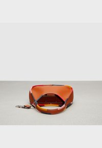 Pieni ruskea nahkalaukku, jossa on monivärisiä tilkkutyydet, pyöreä kahva, avoin yläosa ja kiinnitetty koristeellinen etiketti harmaalla pinnalla.