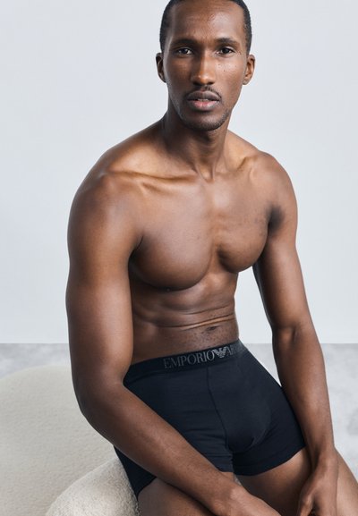 Crne Emporio Armani bokserice s brendiranim pojasom koje nosi muškarac bez majice, prikazujući glatku kožu i definirane mišiće, s minimalnom pozadinom.