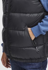 Schwarze Daunenweste mit gestepptem Muster, seitlichem Reißverschlusstaschen und hohem Kragen, getragen über einem blauen Sweatshirt.