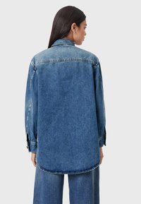 Sinine teksajope, millel on lõdvestunud istuvus, nööbitavad mansetid ja kõverdatud ääris. Iseloomulik tuhmunud tekstuur nähtavate õmblusdetailidega.