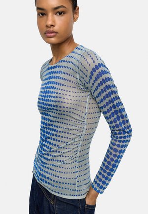 Kobieta w dopasowanej koszulce z długim rękawem, wzór z niebieskich okręgów na szarym tle, w ciemnych dżinsach, stojąca z jedną ręką w kieszeni.