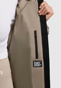 Beige jacket with a smooth texture, featuring a black zipper pocket and a black accent along the edge. Branding patch on the interior.