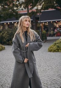 Długi, szary wełniany płaszcz z szerokimi rękawami i paskiem. Posiada teksturowane wykończenie i sięga do kolan. Nosi się go na czarną bluzkę.
