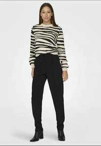 Mulher vestindo um suéter listrado em preto e branco, calças pretas de cintura alta e botas pretas até o tornozelo, de pé contra um fundo simples.