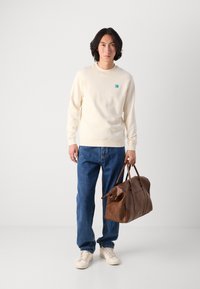 Crème sweatshirt met een klein turquoise logo, blauwe jeans, beige sneakers en een grote leren handtas in bruin. Gladde textuur overal.