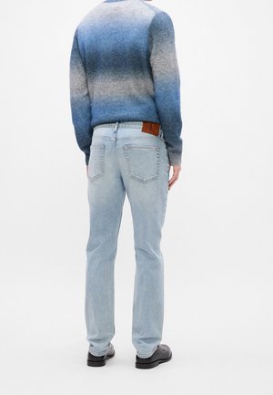 Mann trägt hellblaue Jeans und einen blau verlaufenden Pullover mit langen Ärmeln, steht mit dem Rücken zum Betrachter vor weißem Hintergrund.