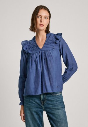 Blå bomuldsbluse med ruffles på kraven og ærmerne, der har plisseringer foran og samlede manchetter. Bæres med jeans.