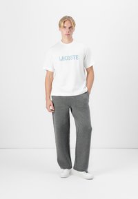 Biele bavlnené tričko s logom "LACOSTE" v modrej a zelenej farbe, skombinované s svetlosivými pletenými teplákmi a bielymi teniskami. Jednoduchý dizajn.