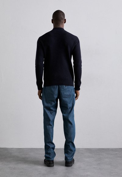 Pulover tricotat cu rețea navală, asortat cu pantaloni din denim bleu deschis, cu croială lejeră; ambele purtate cu încălțăminte neagră, văzute din spate pe un fundal neutru.