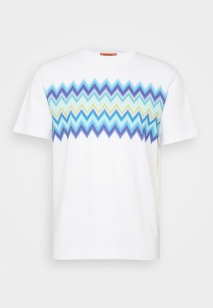 T-shirt en coton blanc avec un motif en zigzag bleu, vert et bleu clair sur la poitrine. Encolure ronde et manches courtes.