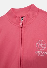 Pink zip-up jacket made of soft fabric, featuring a high collar, branded zipper pull, and embroidered logo on the chest.