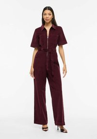 Tuta in velluto a coste color bordeaux con maniche corte, zip frontale, due tasche sul petto, vita annodata e pantaloni a gamba larga. Tessuto liscio.