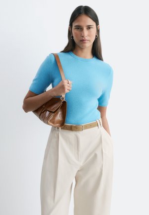 Mujer con cabello largo y oscuro que lleva una camiseta azul de manga corta y pantalones beige de cintura alta, sosteniendo un bolso de cuero marrón al hombro.