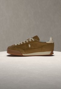 Baskets en daim marron avec des lacets blancs, une bordure blanche de la semelle et un petit logo blanc de joueur de polo sur le côté, présentées sur une surface claire.