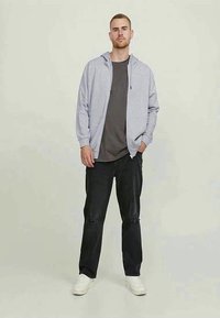 Siva zip-up kapuca, teksturirana tkanina, preko tamne majice, u kombinaciji s crnim izbrazdanim trapericama i bijelim tenisicama. Stojeći protiv svijetle pozadine.