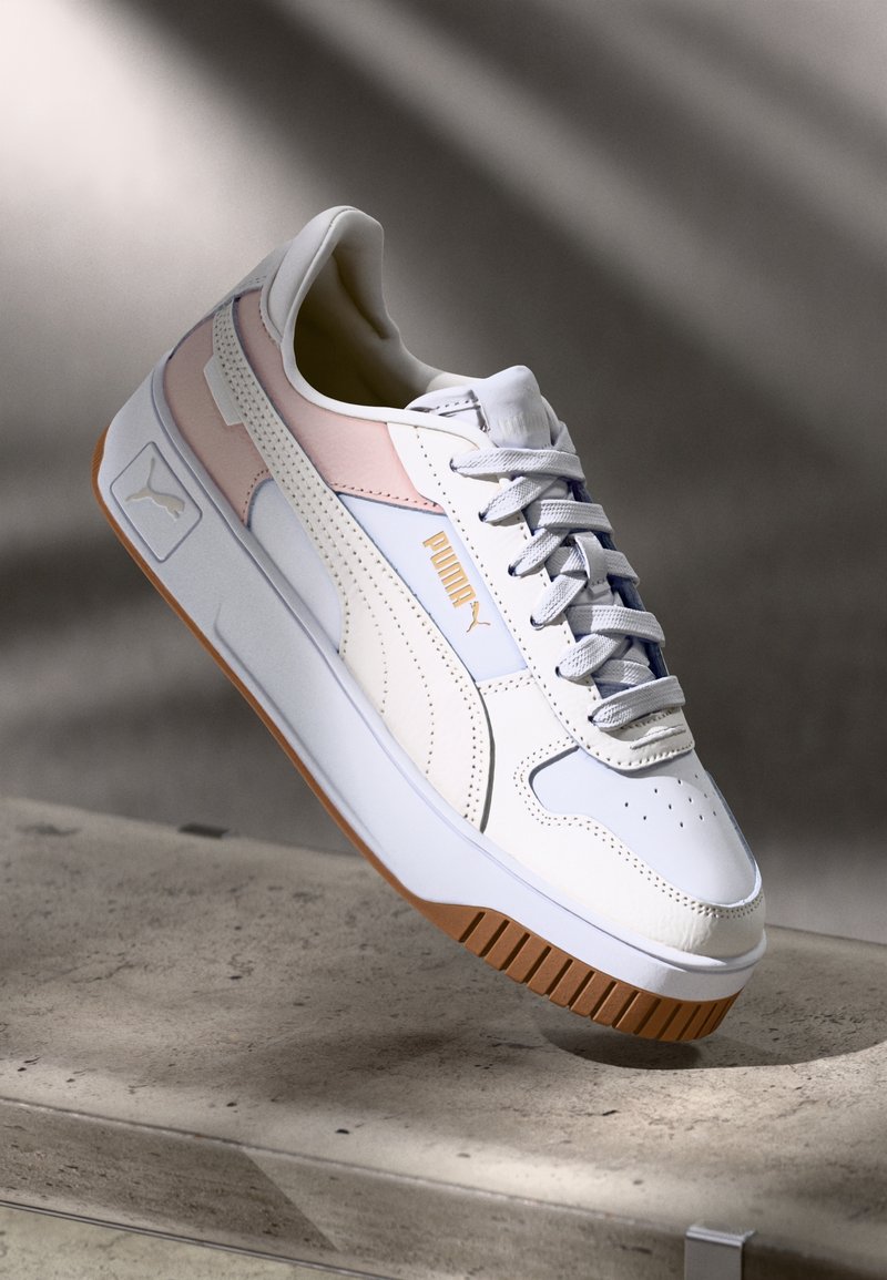 Bílé a světle růžové tenisky Puma s gumovou podešví a tkaničkami vystavené na betonovém povrchu s jemným stínem v pozadí.