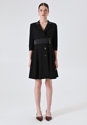 Robe noire avec un col et des manches trois-quarts. Elle présente une taille cintrée avec trois boutons décoratifs et une jupe évasée.
