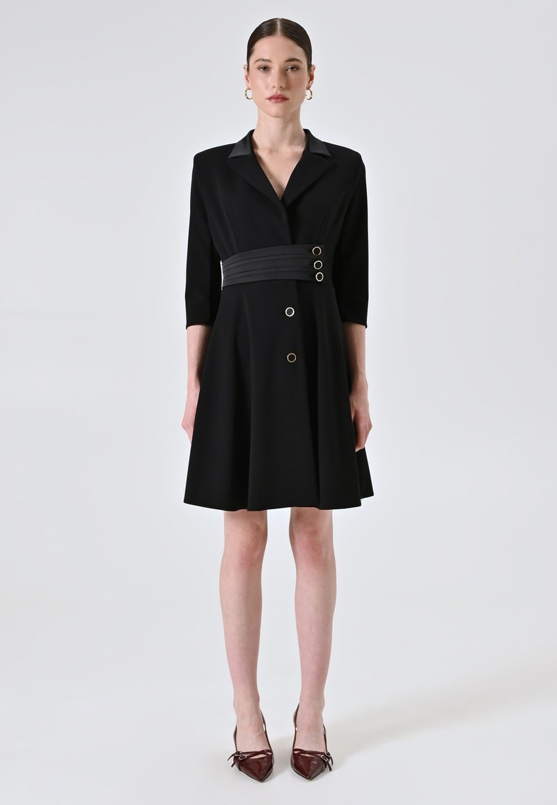 Zwarte jurk met een kraaghalslijn en driekwartmouwen. Heeft een getailleerde taille met drie decoratieve knopen en een uitgerokken rok.