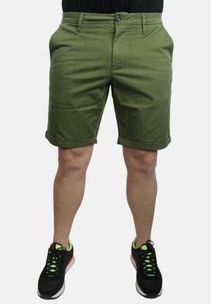 Pantaloni corti verde oliva con chiusura a bottone, tasche laterali e risvolti, realizzati in un tessuto morbido, abbinati a scarpe da ginnastica nere.