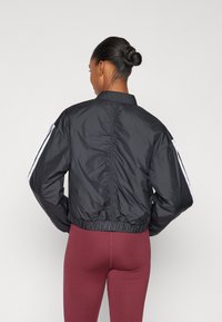 Črna skrajšana jakna iz gladkega, lahkega materiala, ki ima bele črte na ramenih in zbrano obrobo.