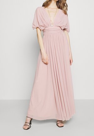 Robe maxi en tissu rose doux avec un décolleté en V profond, manches courtes drapées et jupe plissée. Associée à des sandales noires à fines lanières.
