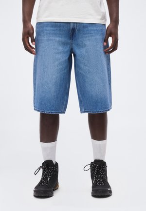 Persoană purtând pantaloni scurți din denim albastru până la genunchi, șosete albe până la gleznă, bocanci negri cu șireturi și un tricou alb, stând pe un fundal deschis.