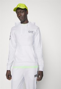 Felpa bianca con cappuccio e cordini, logo EA7 sul petto e sulle maniche. Abbinata a dettagli verde fluo nel colletto e nei polsini. Finitura in tessuto morbido.