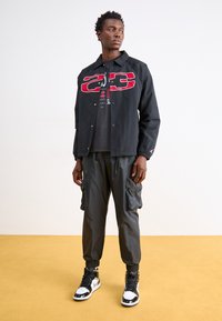 Μαύρο ελαφρύ μπουφάν με κόκκινο γραφικό, σε συνδυασμό με σκούρο cargo παντελόνι και μαύρα ψηλά sneakers με λευκές και ασημί λεπτομέρειες.