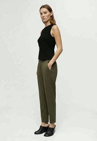 Zwarte mouwloze top, olijfgroene vormgegeven broek en zwarte instap schoenen. Model staat zijwaarts, toont een getailleerd silhouet en zakken.