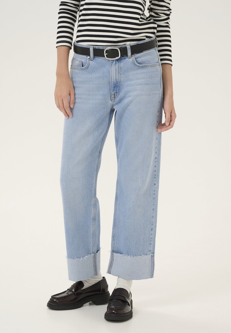 Jeans de tiro alto de pierna ancha en azul claro con dobladillo deshilachado, con cinco bolsillos y un cinturón negro. La modelo lleva mocasines oscuros y calcetas grises.