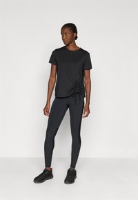 T-shirt noir à manches courtes avec fronces sur les côtés, associé à un legging foncé et des chaussures de sport noires. Tissu lisse avec une coupe décontractée.