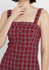 Abito a quadri rosso con scollo quadrato, spalline sottili e bottoni di legno lungo la parte anteriore. Il tessuto ha una consistenza liscia con un motivo a scacchi.