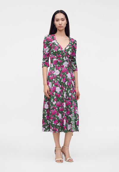 Γυναίκα με πράσινο floral midi φόρεμα τύπου wrap με ροζ και λευκά λουλούδια και μπεζ ψηλοτάκουνα σανδάλια, στέκεται μπροστά σε απλό φόντο.