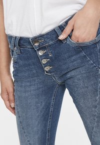 Vaqueros de mezclilla azul con un diseño de talle alto, cierre frontal de cinco botones y detalles de costura visibles. Bolsillo lateral visible en la izquierda.