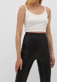 Top corto bianco a coste con spalline larghe, abbinato a pantaloni in finta pelle neri a vita alta, con una texture liscia e lucida.