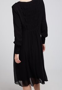 Czarna sukienka z długimi rękawami, teksturowanym gorsetem z marszczeniami i zwiewną spódnicą. Lekki materiał z przezroczystym dołem.