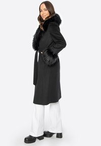 Schwarzer Wollmantel mit einem Kunstfellkragen und -manschetten, tailliert, mit geradem Schnitt und langen Ärmeln.