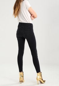 Černé úzké džíny s vysokým pasem a hladkou strukturou, vybavené zadními kapsami, kombinované se lesklými zlatými kotníkovými botami.