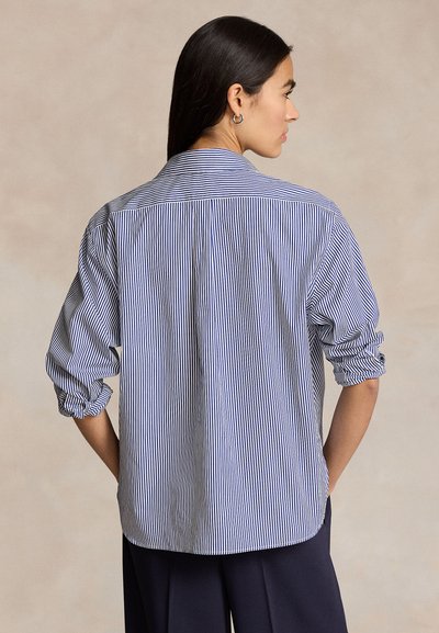 Žena s dlhými tmavými vlasmi, ktorá nosí modro-bielu vertikálne pruhovanú košeľu s vyhrnutými rukávmi, otočená chrbtom k fotoaparátu.