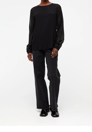 Personne portant un chemisier noir à manches longues avec des manches transparentes à motifs, un jean noir ample avec des ourlets effilochés et des chaussures noires brillantes.