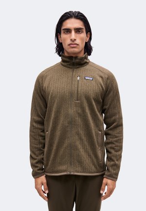 Hombre con chaqueta de forro polar verde oliva con cremallera completa, cuello alto y bolsillo con cremallera en el pecho, de pie delante de un fondo liso.