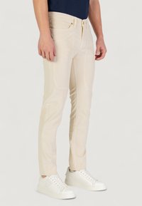 Pantaloni beige slim-fit in velluto a coste, dotati di quattro tasche, chiusura con zip e bottone, e una superficie texturizzata, abbinati a sneaker bianche.