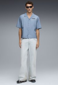 Světle modrá košile s krátkým rukávem má na sobě logo nášivku, je kombinována s volnými světlými džíny a černými teniskami. Matná textura po celé ploše.