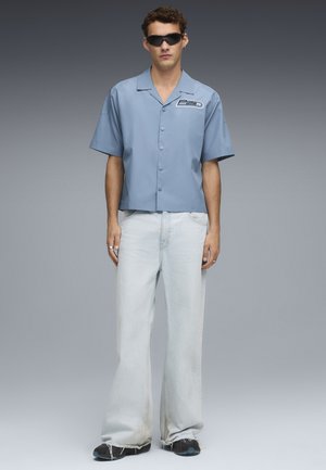 Camisa de manga corta azul claro con un parche de logo, combinada con unos vaqueros de mezclilla claros de corte holgado y zapatillas negras. Textura mate en toda la prenda.