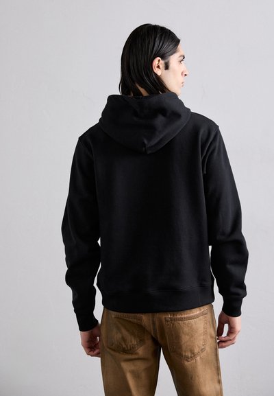 Un sweat à capuche noir avec une texture douce, des poignets et un ourlet côtelés. Porté avec un pantalon marron clair, montrant une vue arrière décontractée.