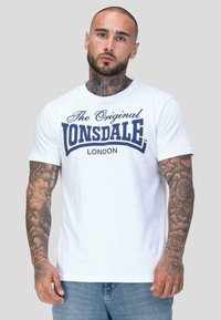 Mees, kellel on tätoveeringud kaelal ja kätes, kannab valget Lonsdale London T-särki ja siniseid teksaseid, seistes ühetaolise tausta ees.