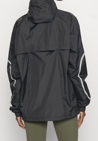 Chaqueta negra impermeable con capucha, que presenta detalles reflectantes, un corte relajado y puños elásticos. Textura suave con un diseño minimalista.