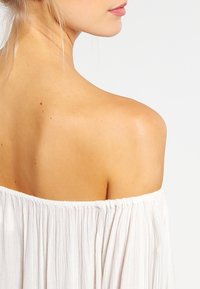 Off-shoulder vit blus med textured tyg och samlad halsringning. Slät hudton synlig på axel och rygg.