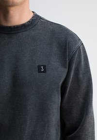 Donkergrijze sweatshirt van een gestructureerde stof, met een geribbelde halslijn en een klein zwart vierkant patch met een witte letter "J" aan de voorkant.
